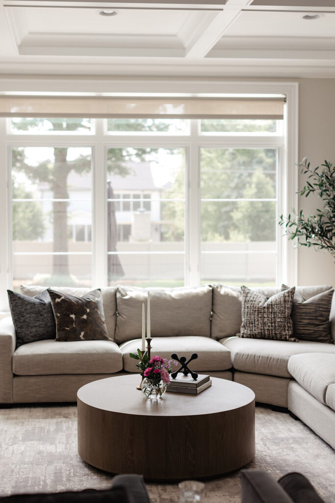 A modern living room with a large beige sectional sofa, patterned pillows, a round wooden coffee table with decor, and large windows overlooking a green yard.