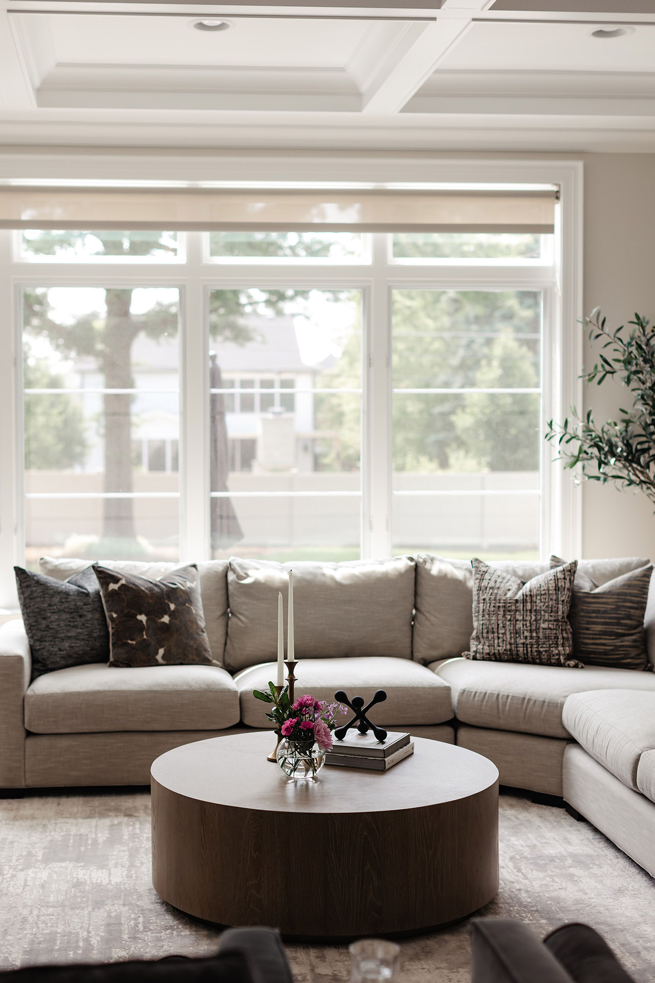 A modern living room with a large beige sectional sofa, patterned pillows, a round wooden coffee table with decor, and large windows overlooking a green yard.