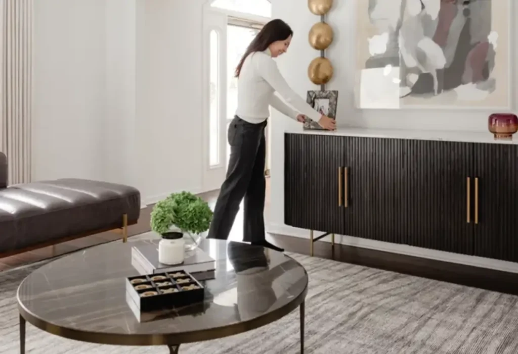 A woman arranges decor on a black sideboard in a modern living room with a round coffee table, abstract wall art, and neutral furnishings.