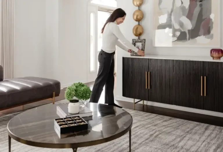 A woman arranges decor on a black sideboard in a modern living room with a round coffee table, abstract wall art, and neutral furnishings.