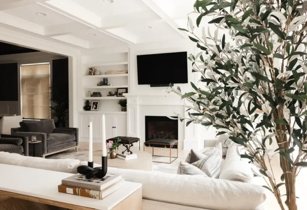 Modern living room with white walls, built-in shelves, a fireplace, mounted TV, light sofa, dark armchairs, large potted plant, and decor items on a wooden console table.