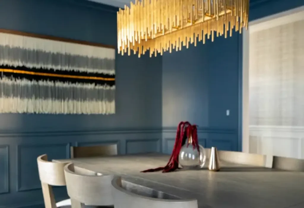 A modern dining room with a gray table, light wood chairs, a glass vase with red branches, a gold geometric chandelier, blue paneled walls, and abstract wall art.
