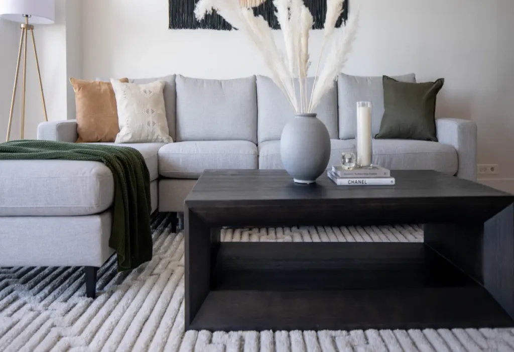 A modern living room with a light gray sectional sofa, neutral throw pillows, a dark wood coffee table, a vase with pampas grass, and a textured area rug.