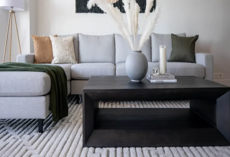 A modern living room with a light gray sectional sofa, neutral throw pillows, a dark wood coffee table, a vase with pampas grass, and a textured area rug.