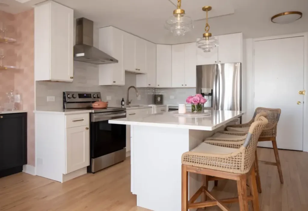 Modern kitchen with white cabinets, stainless steel appliances, a center island with two woven stools, and pink flowers in a vase on the countertop.