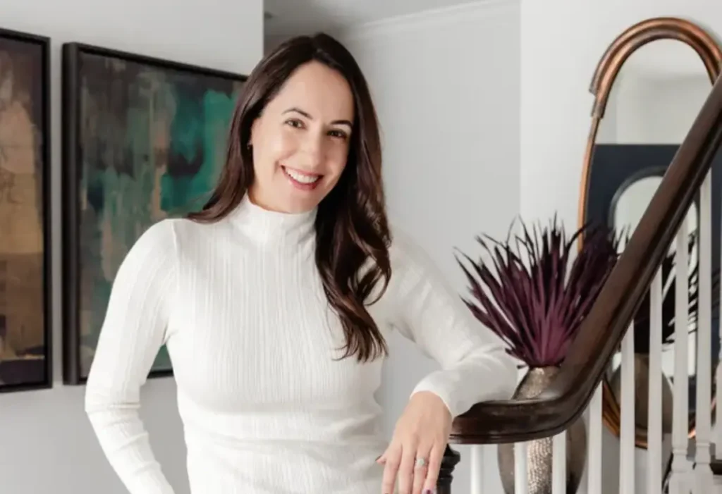 A woman in a white long-sleeve top stands indoors by a staircase, smiling at the camera. There is a purple plant and artwork in the background.