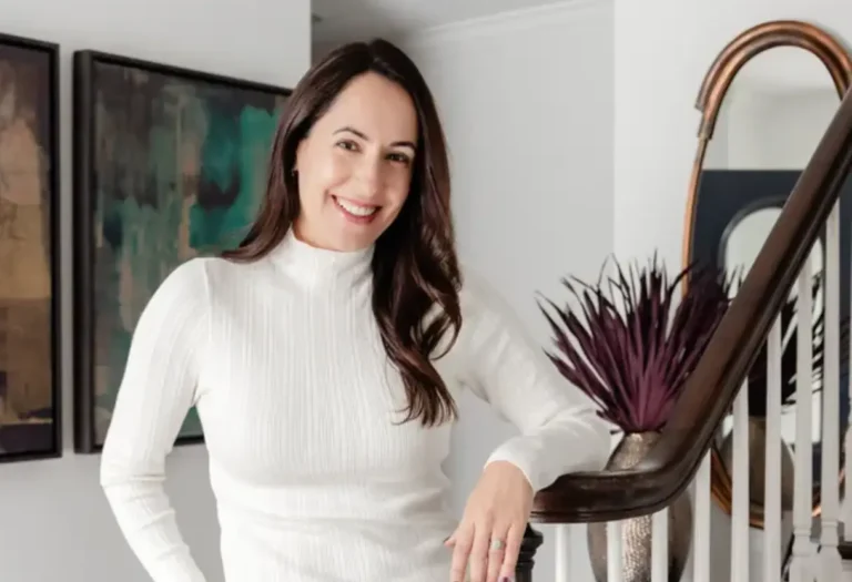 A woman in a white long-sleeve top stands indoors by a staircase, smiling at the camera. There is a purple plant and artwork in the background.