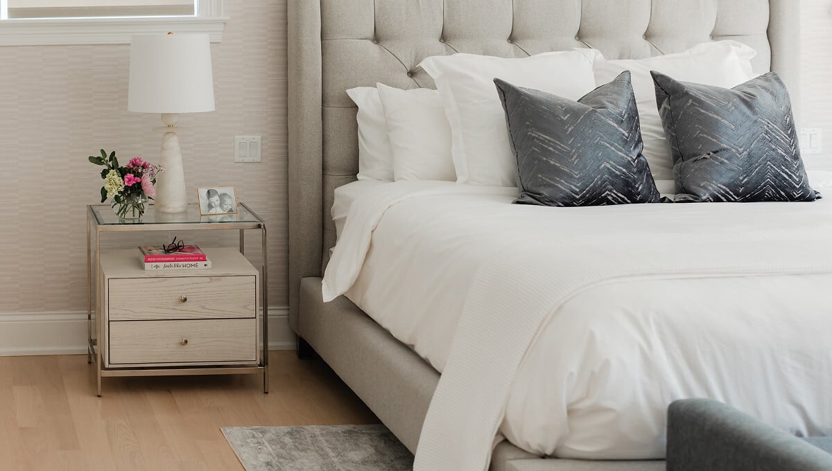 Primary bedroom in an Elmhurst, IL home with a tufted upholstered headboard, layered neutral bedding and a mirrored nightstand, designed by Dara Agruss Design