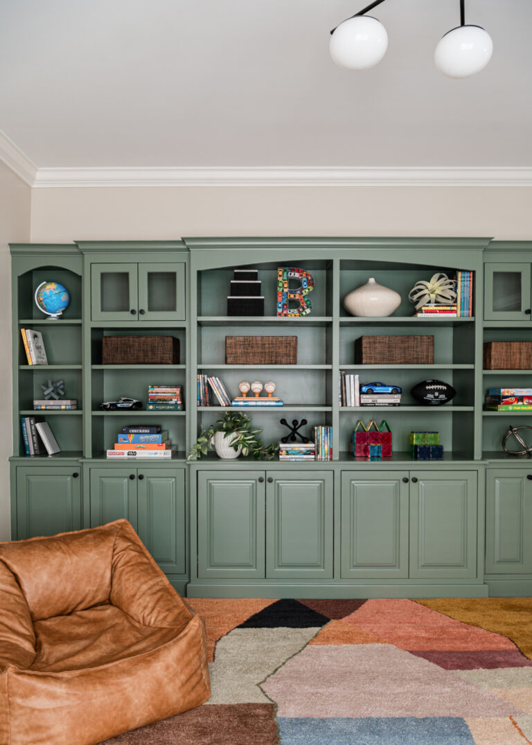 Green built-in shelves with books, toys, and decor items, a tan bean bag chair, and a colorful geometric rug in a well-lit room.
