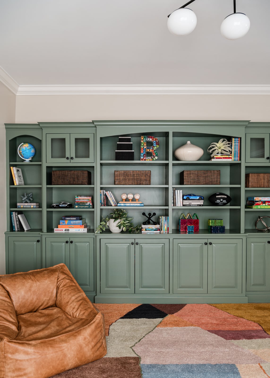The same playroom built-ins after renovation by Dara Agruss Design, painted sage green with styled open shelving, white oak floors and a striped wool rug