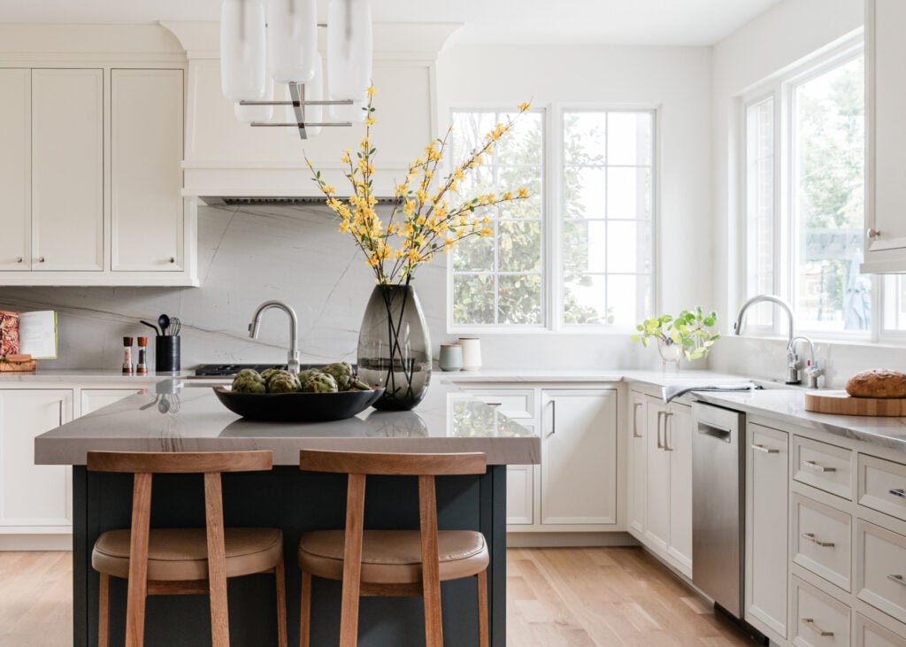 Modern kitchen with white cabinets, a large island with two wooden stools, a vase of yellow flowers, and natural light coming through two large windows.