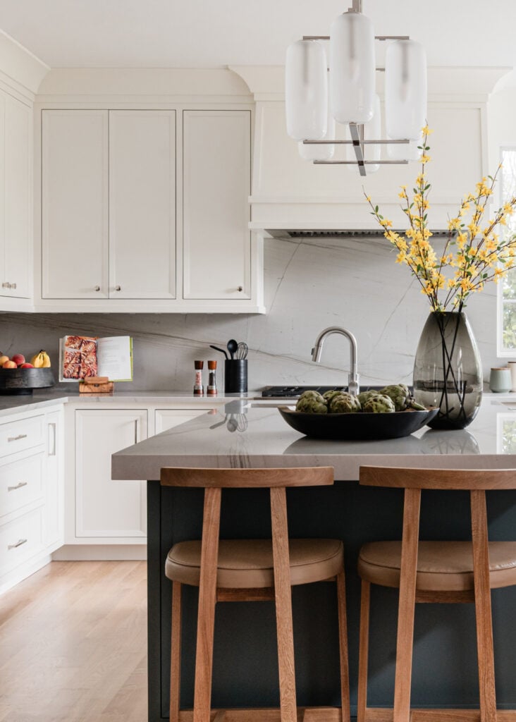 Modern kitchen with white cabinets, gray countertops, a black island with two wooden stools, a pendant light, and decorative vases with yellow flowers on the counter.