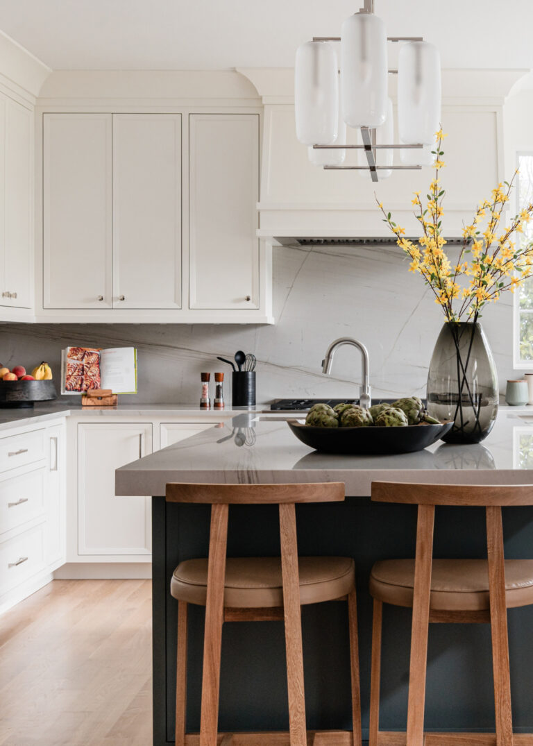 Modern kitchen with white cabinets, gray countertops, a black island with two wooden stools, a pendant light, and decorative vases with yellow flowers on the counter.