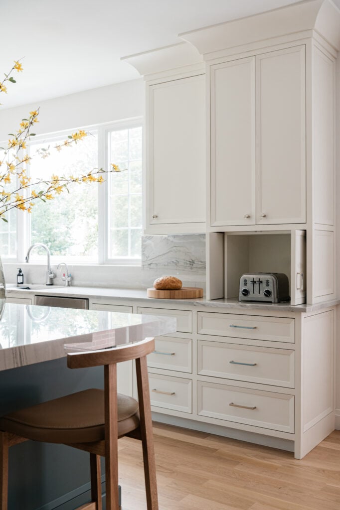 Modern kitchen with white cabinets, marble backsplash, a loaf of bread on a cutting board, a toaster, and a large window letting in natural light.