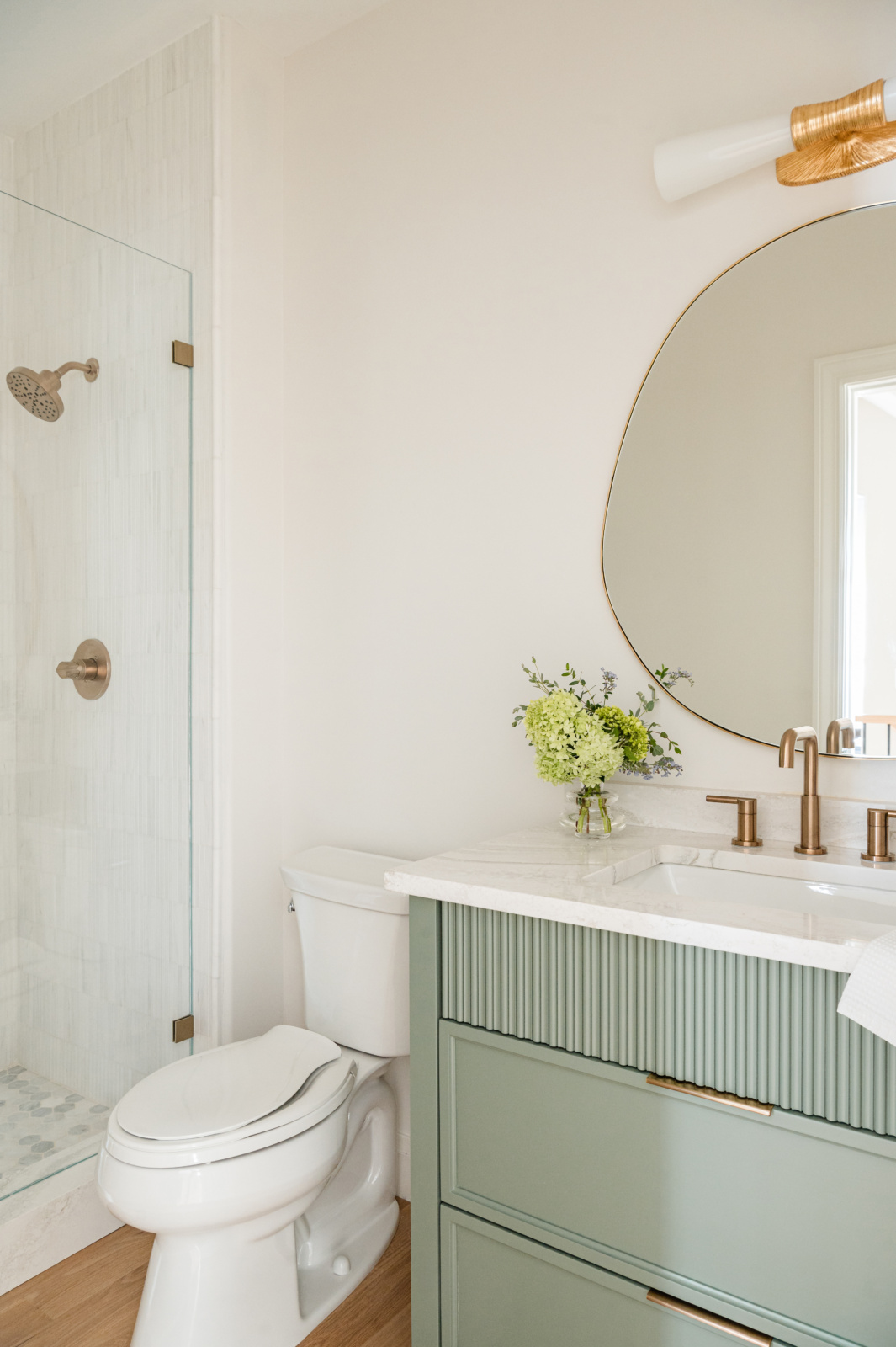 Bathroom with a glass shower, white toilet, green vanity with marble countertop, brass fixtures, round mirror, and a small vase of flowers.
