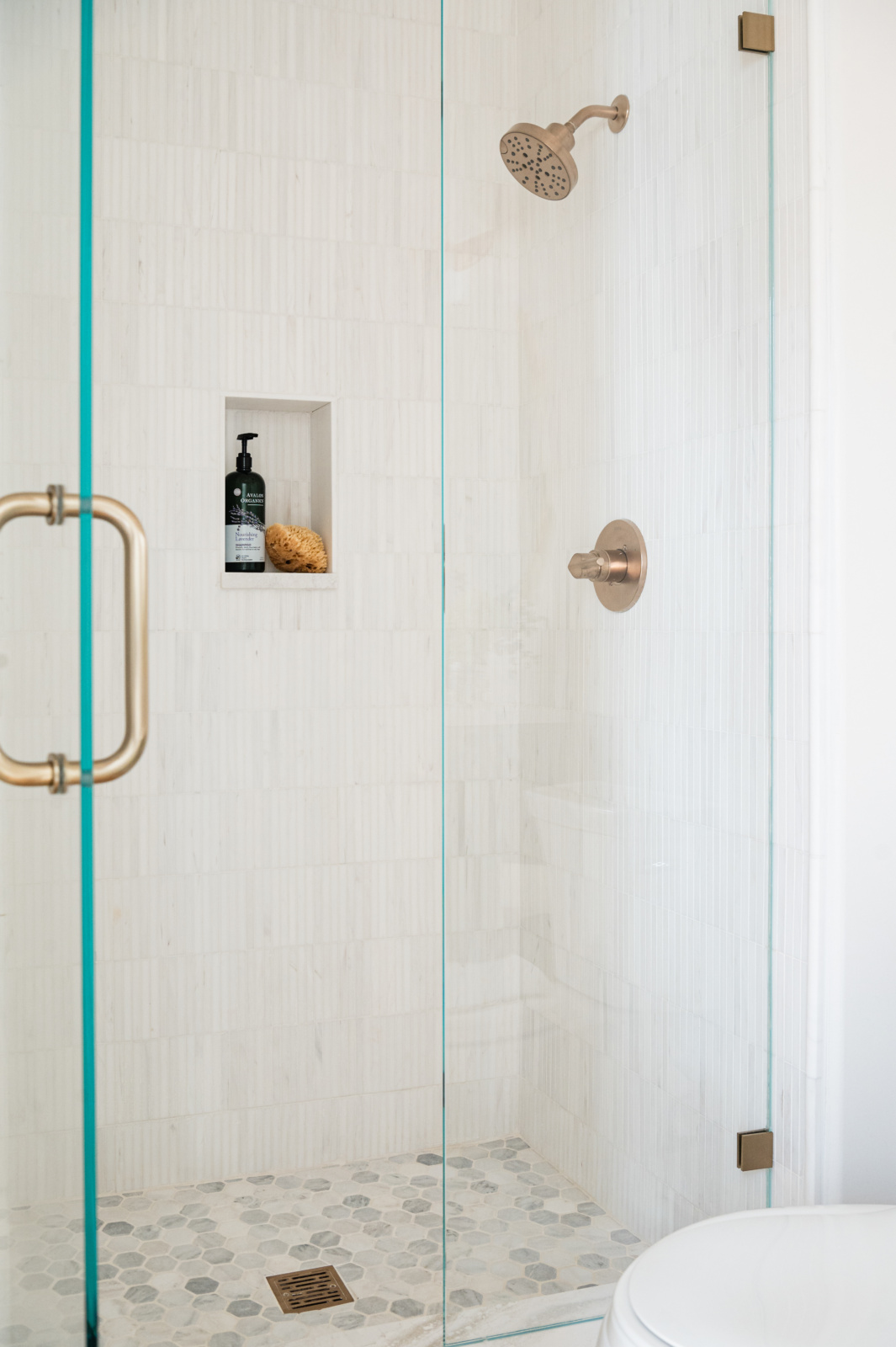 Glass-enclosed shower with white tile walls, built-in shelf holding a black soap dispenser and sponge, and bronze fixtures including a showerhead and handle.