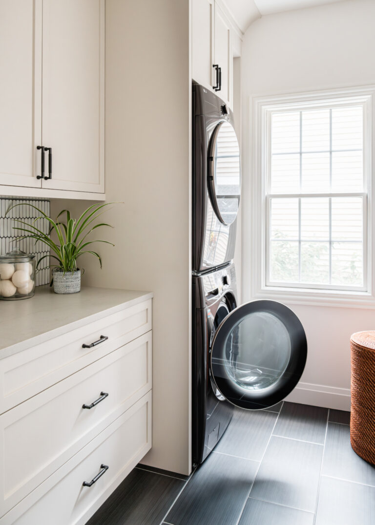 Stacked black washer and dryer with the washer door open in a bright laundry room with white cabinets, a plant, and a wicker basket by a large window.
