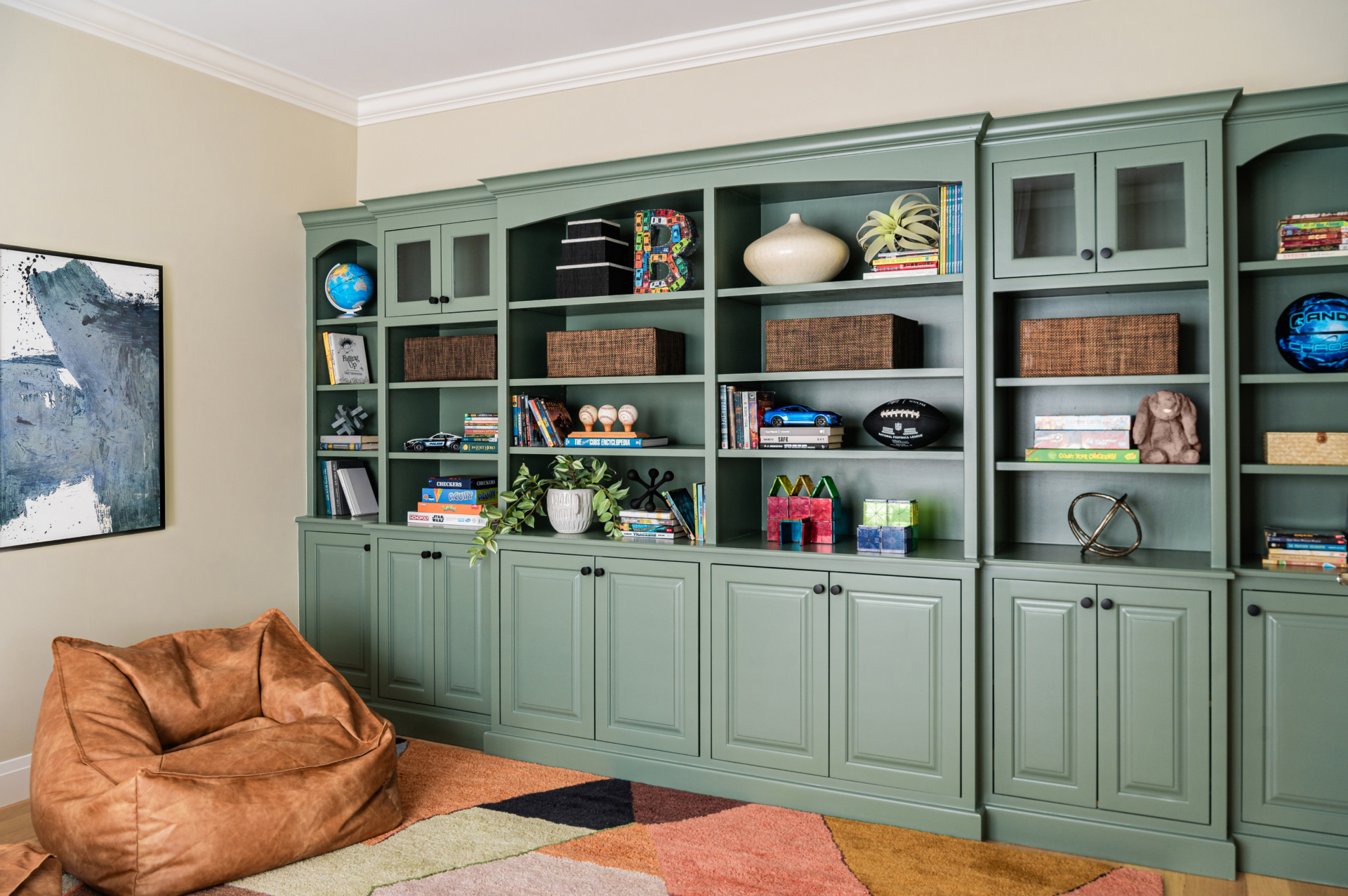 Green built-in shelving unit filled with books, baskets, and decorative items, next to a brown bean bag chair and colorful rug in a cozy room.