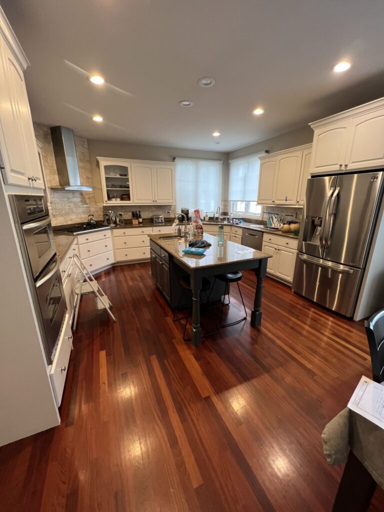 Modern kitchen with white cabinets, stainless steel appliances, a central island with stools, and hardwood floors. Countertops and island have various items placed on them.