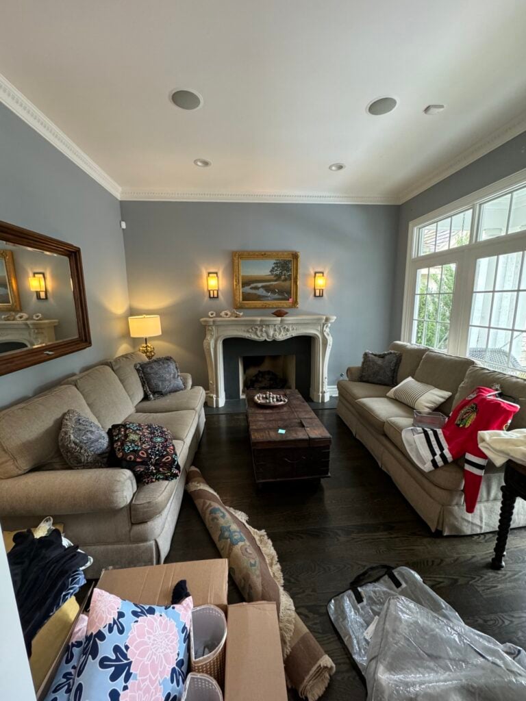 A living room with two beige sofas facing a white fireplace, a wooden coffee table, large windows, and various items, including clothing and boxes, scattered around.