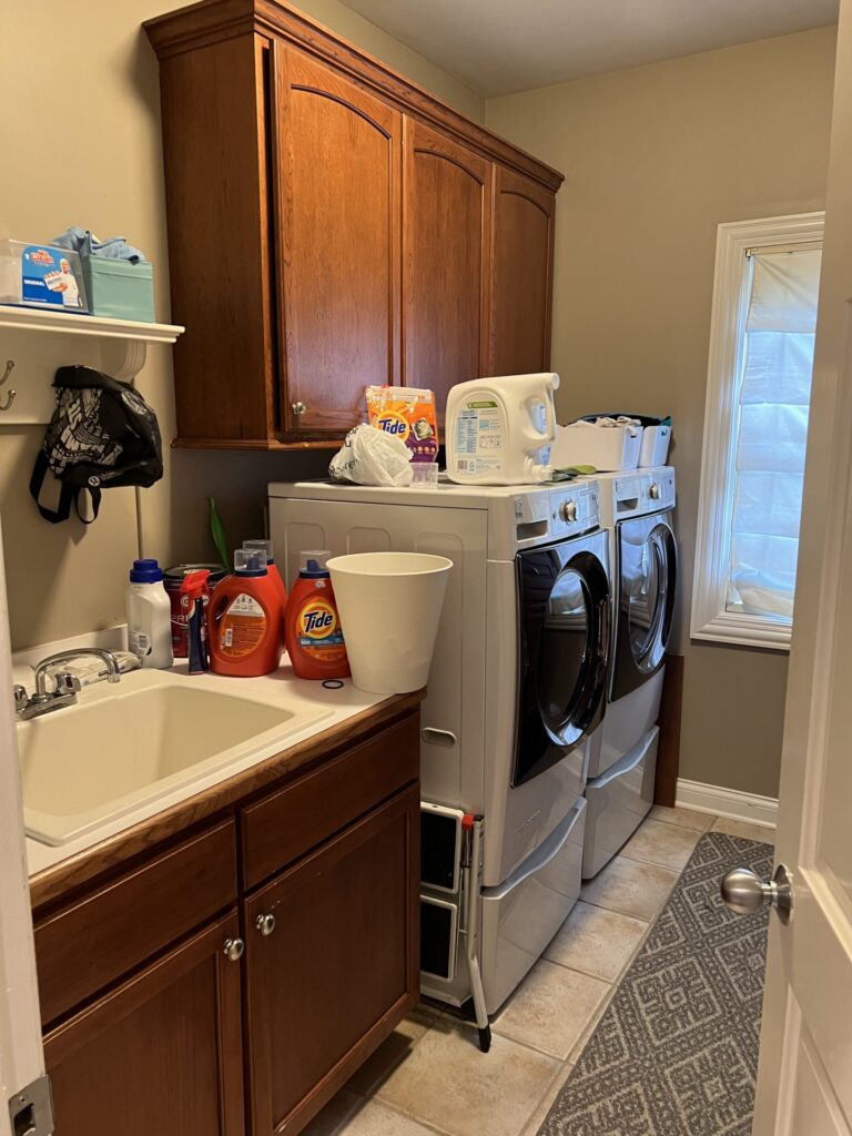 Laundry room with a sink, wooden cabinets, stacked washer and dryer, laundry detergent bottles, a laundry basket, and cleaning supplies. A window with a shade is on the right wall.