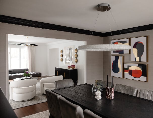 Modern dining room with a dark wood table and gray chairs, abstract art on the wall, and a circular light fixture; living room with white chairs and wall decor visible in the background.