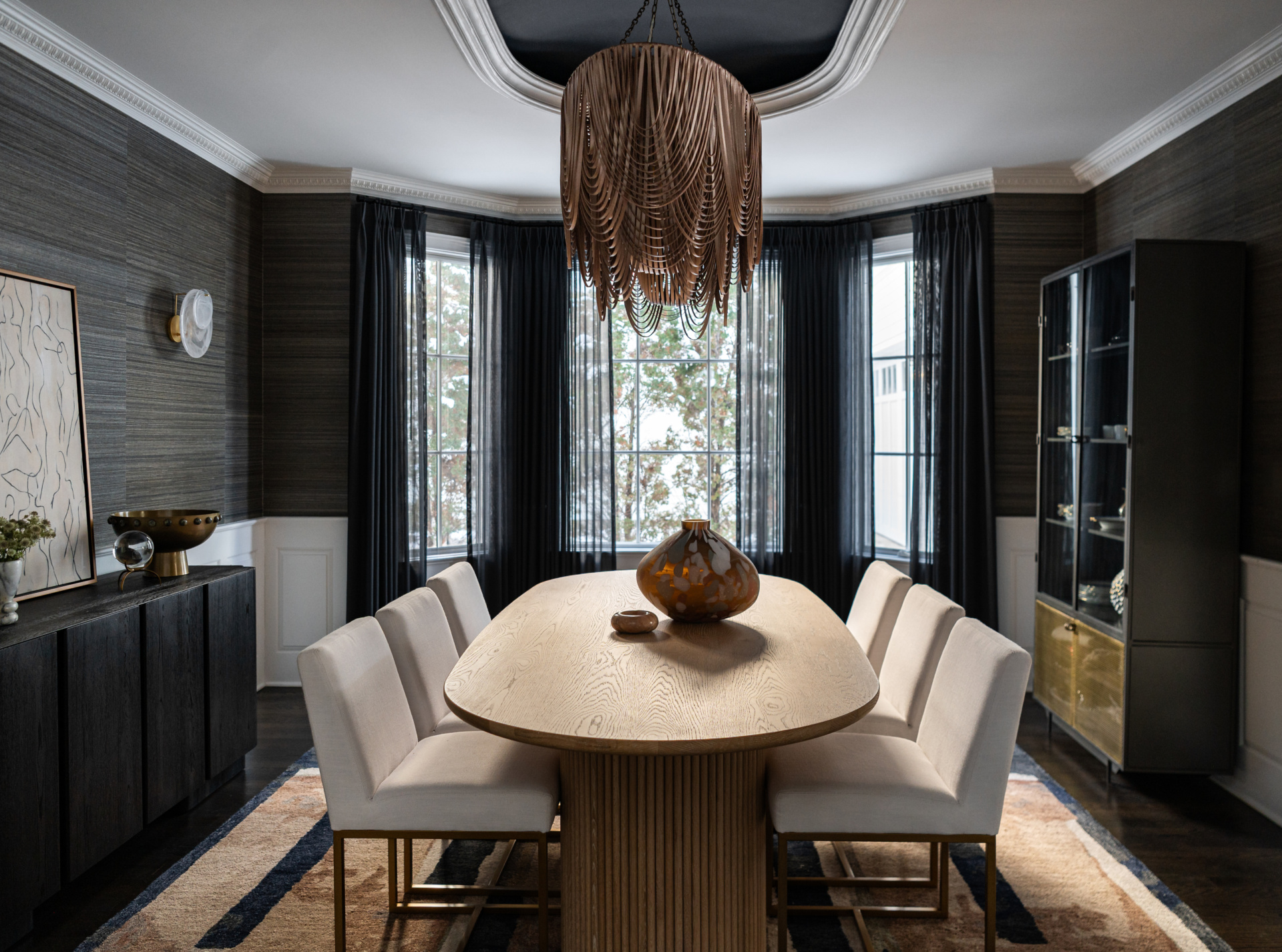 Modern dining room with a wooden oval table, eight white cushioned chairs, a large decorative vase, dark curtains, and a unique chandelier hanging from the ceiling.