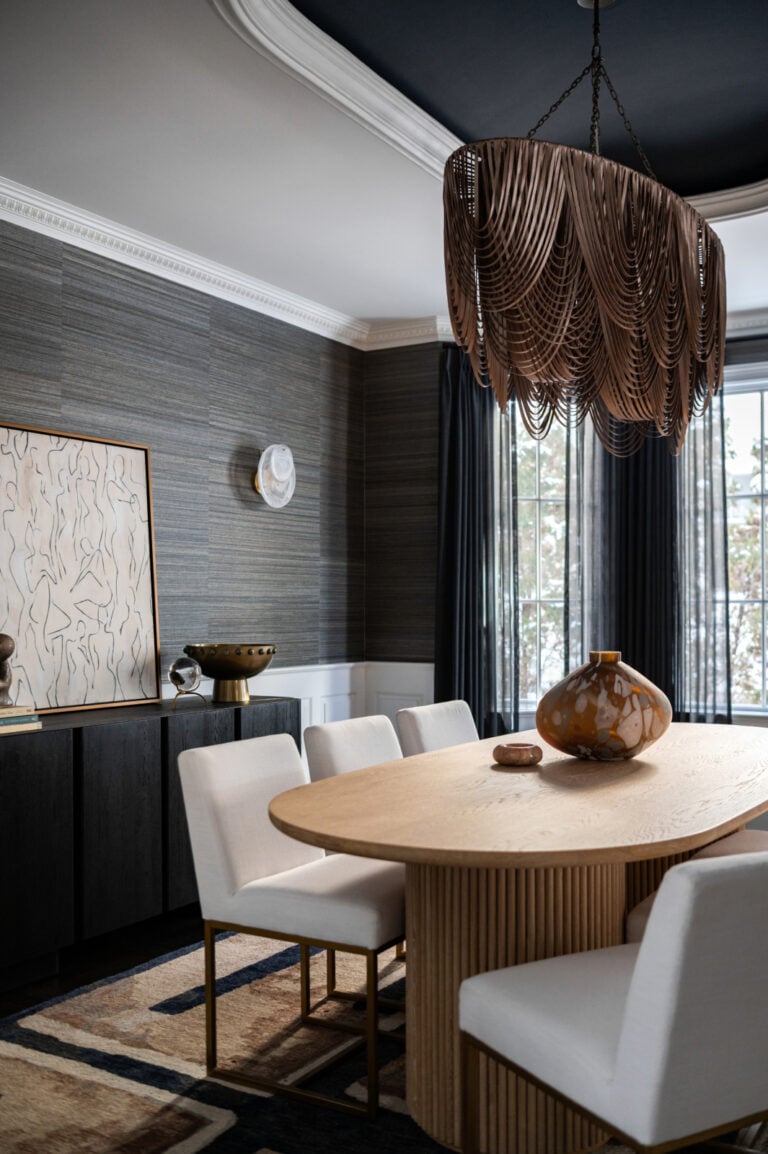 A modern dining room with a wooden oval table, six white chairs, a large decorative chandelier, and contemporary art on the wall. Natural light enters through tall windows with dark curtains.