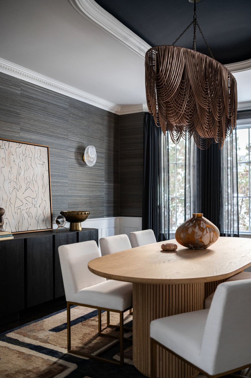 Dining room in Clarendon Hills with textured grasscloth walls, a draped leather chandelier over a light wood oval table with a fluted base, white upholstered chairs, a dark painted ceiling and sheer charcoal drapery at the bay window.