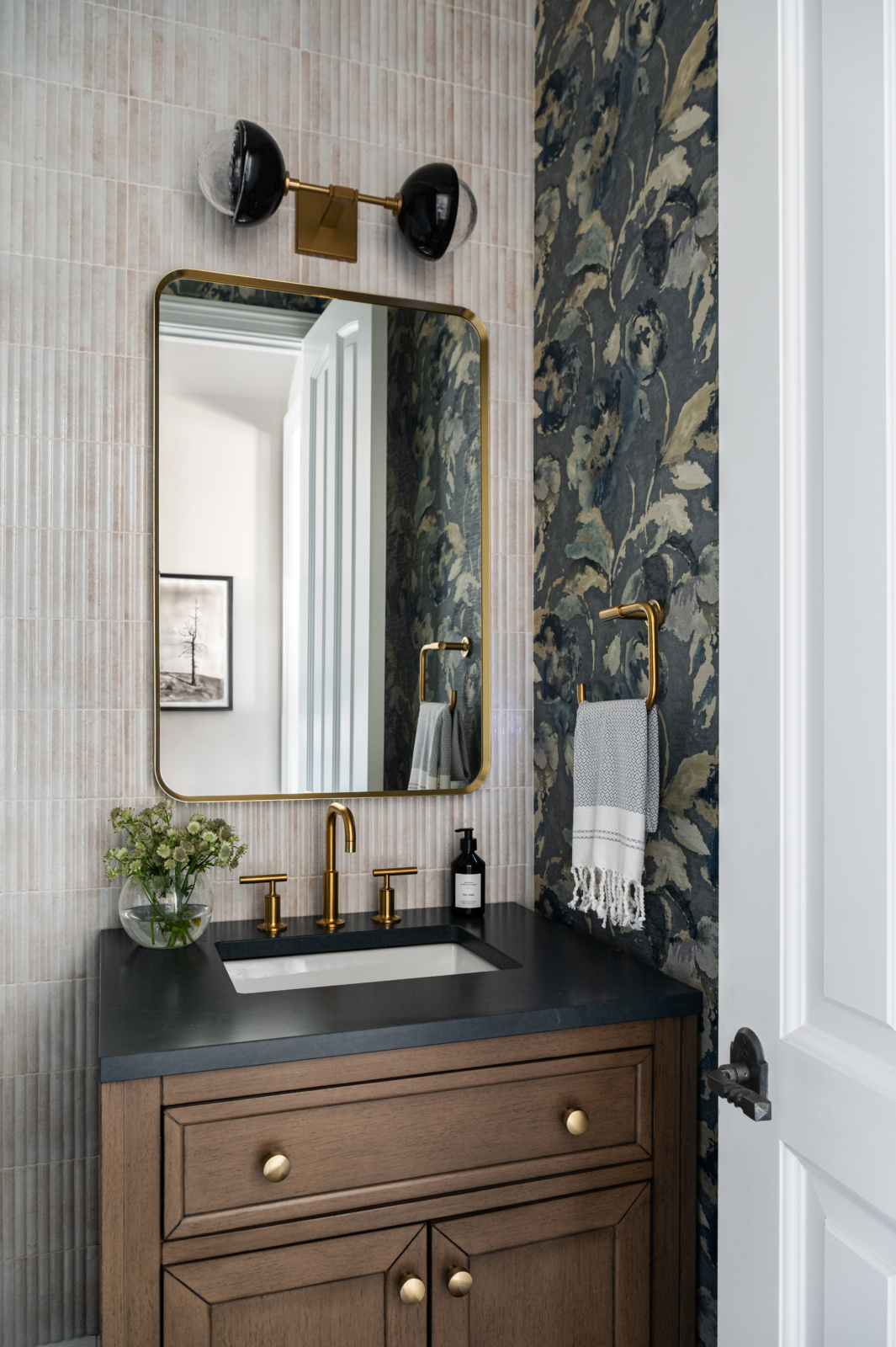 Bathroom vanity with a black countertop, gold fixtures, a rectangular mirror, floral wallpaper, striped wall tiles, and a wall sconce with two black shades above the mirror.