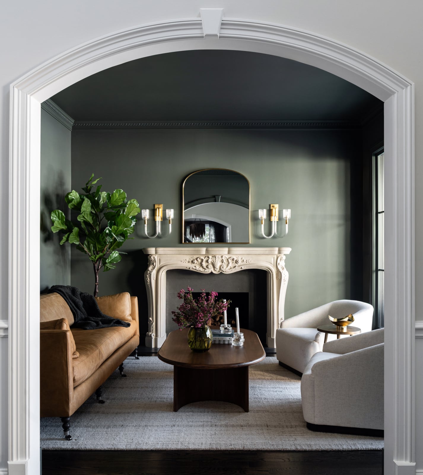 A stylish living room with a brown sofa, two white armchairs, a dark wooden coffee table, ornate fireplace, large mirror, wall sconces, and a potted plant.