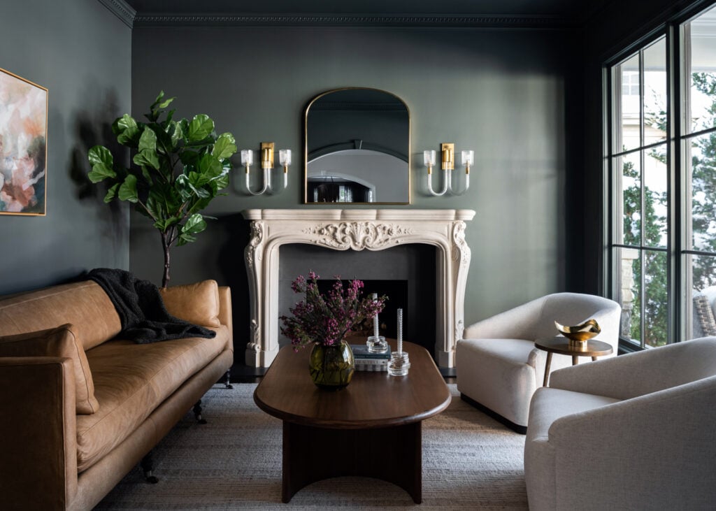 A living room with a brown leather sofa, two white armchairs, a wooden coffee table, a decorative fireplace, wall sconces, and a large indoor plant.