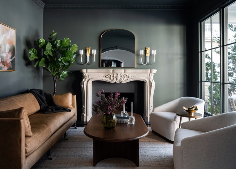 A living room with a brown leather sofa, two white armchairs, a wooden coffee table, a decorative fireplace, wall sconces, and a large indoor plant.