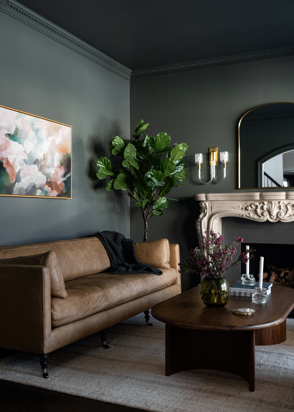 A living room with a tan leather sofa, wood coffee table, ornate fireplace, large potted plant, abstract wall art, and dark gray walls and ceiling.