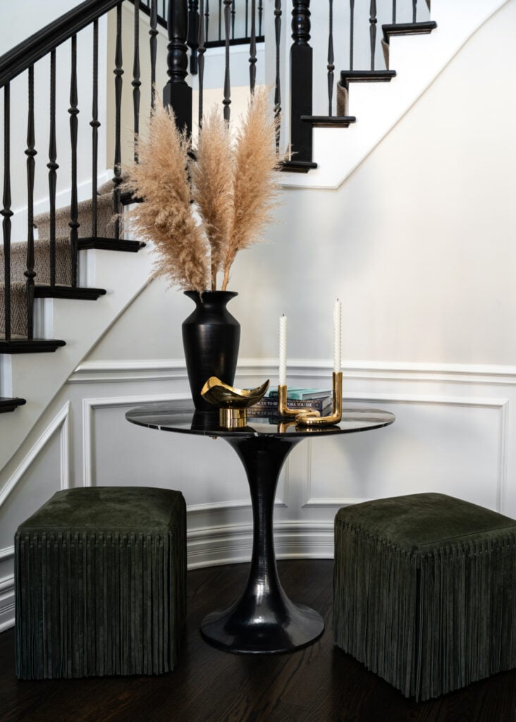 A round black table with gold decor, two green velvet ottomans, and a tall black vase with pampas grass, set against a staircase with black railings and white walls.