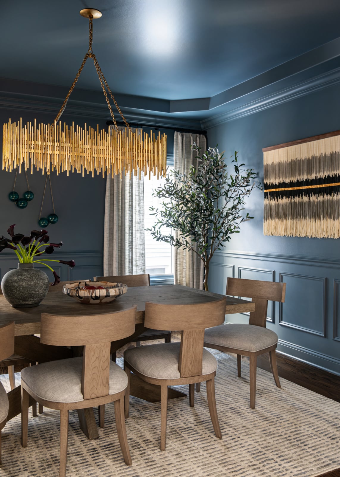A modern dining room with blue walls, a wooden table and chairs, a gold chandelier, a large plant, a vase on the table, and abstract wall art. Natural light enters through a window.