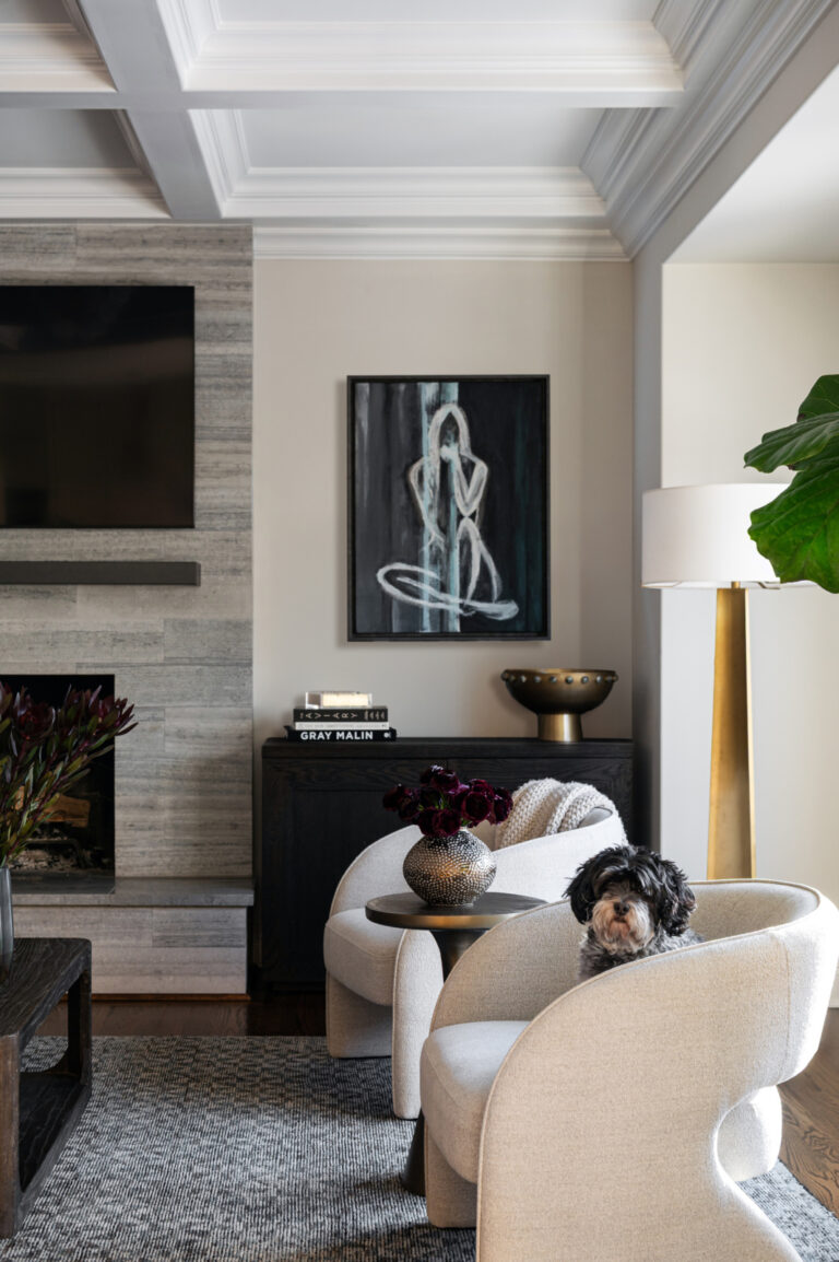 A modern living room with a fireplace, wall-mounted TV, abstract art, two cream armchairs, a small dog sitting on one chair, and a floor lamp beside a large plant.