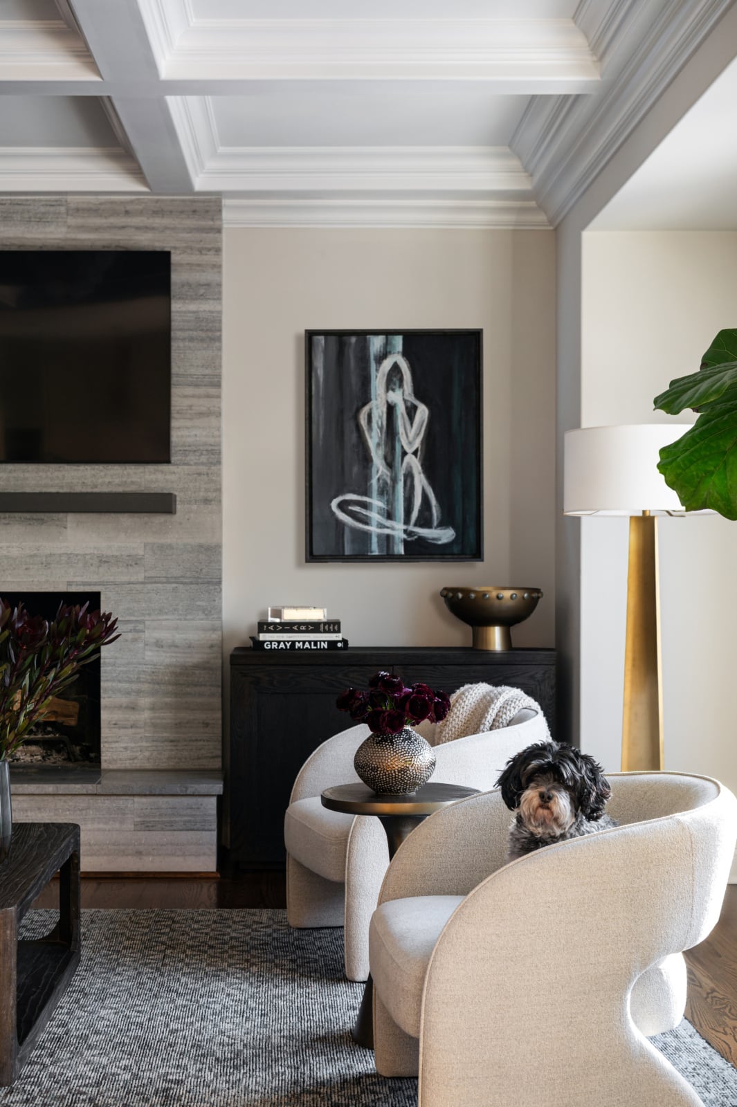 A modern living room with a fireplace, wall-mounted TV, abstract art, two cream armchairs, a small dog sitting on one chair, and a floor lamp beside a large plant.