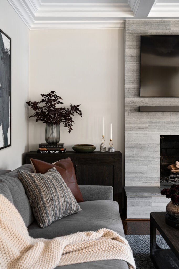 A modern living room with a gray sofa, textured pillows, a cream knit throw, a dark wood console with decor, and a wall-mounted TV above a stone fireplace.