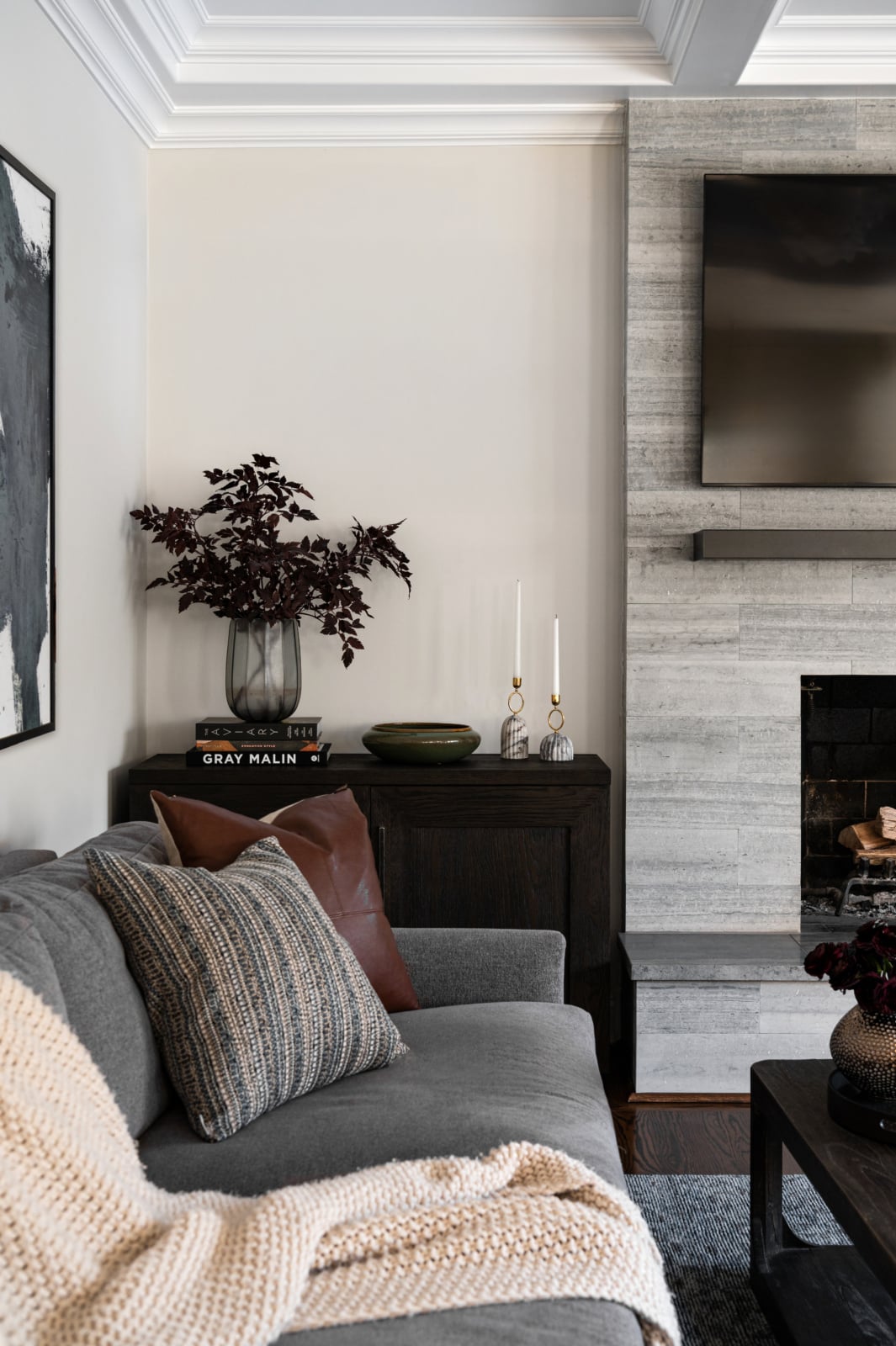 A modern living room with a gray sofa, textured pillows, a cream knit throw, a dark wood console with decor, and a wall-mounted TV above a stone fireplace.
