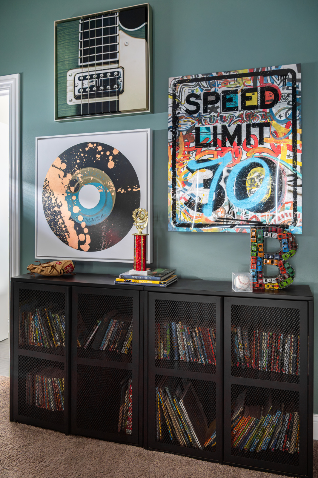 A black bookshelf filled with books sits below wall art featuring a guitar, gold record, speed limit sign, and a decorative letter B. A trophy and toy car are on top of the shelf.