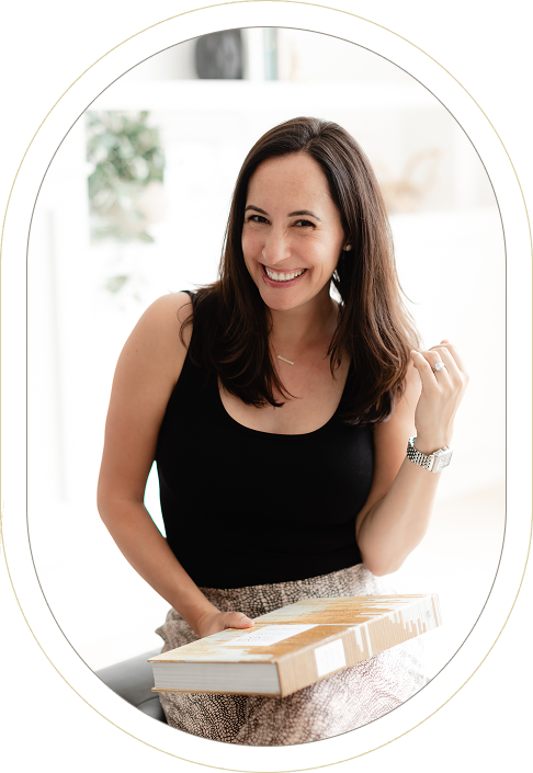 Smiling woman with long brown hair in a black sleeveless top sits holding a closed cardboard box on her lap in a bright indoor setting.