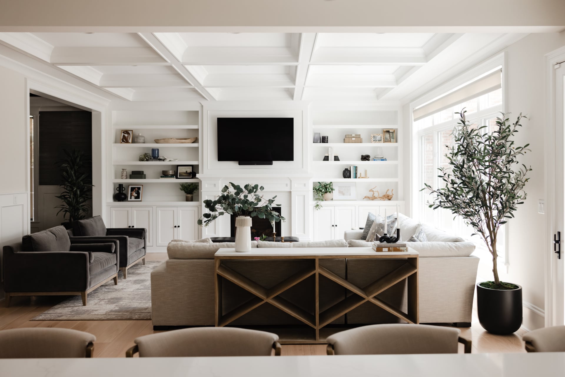 Family room in a newly built home with a coffered ceiling, built-in shelving on either side of the fireplace, a sectional with a wood console behind it, gray velvet armchairs and tall windows.