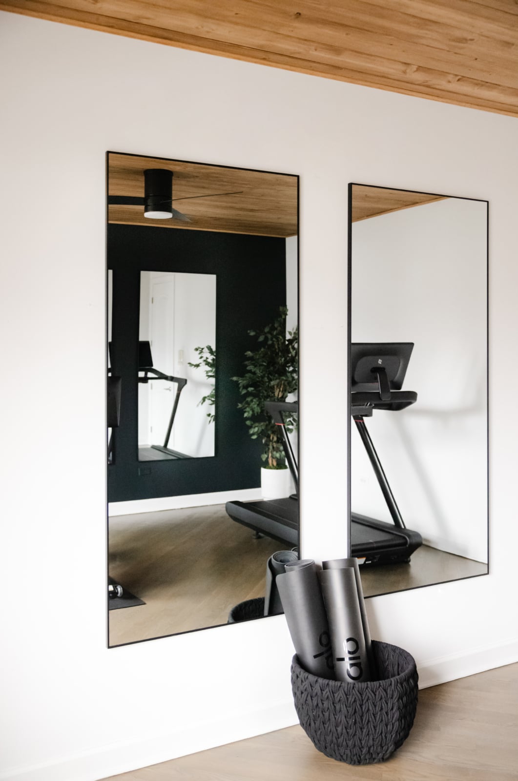 Two wall mirrors reflect a treadmill and a potted plant in a modern, minimalist home gym with light wood flooring and ceiling. A basket with yoga mats sits in the foreground.
