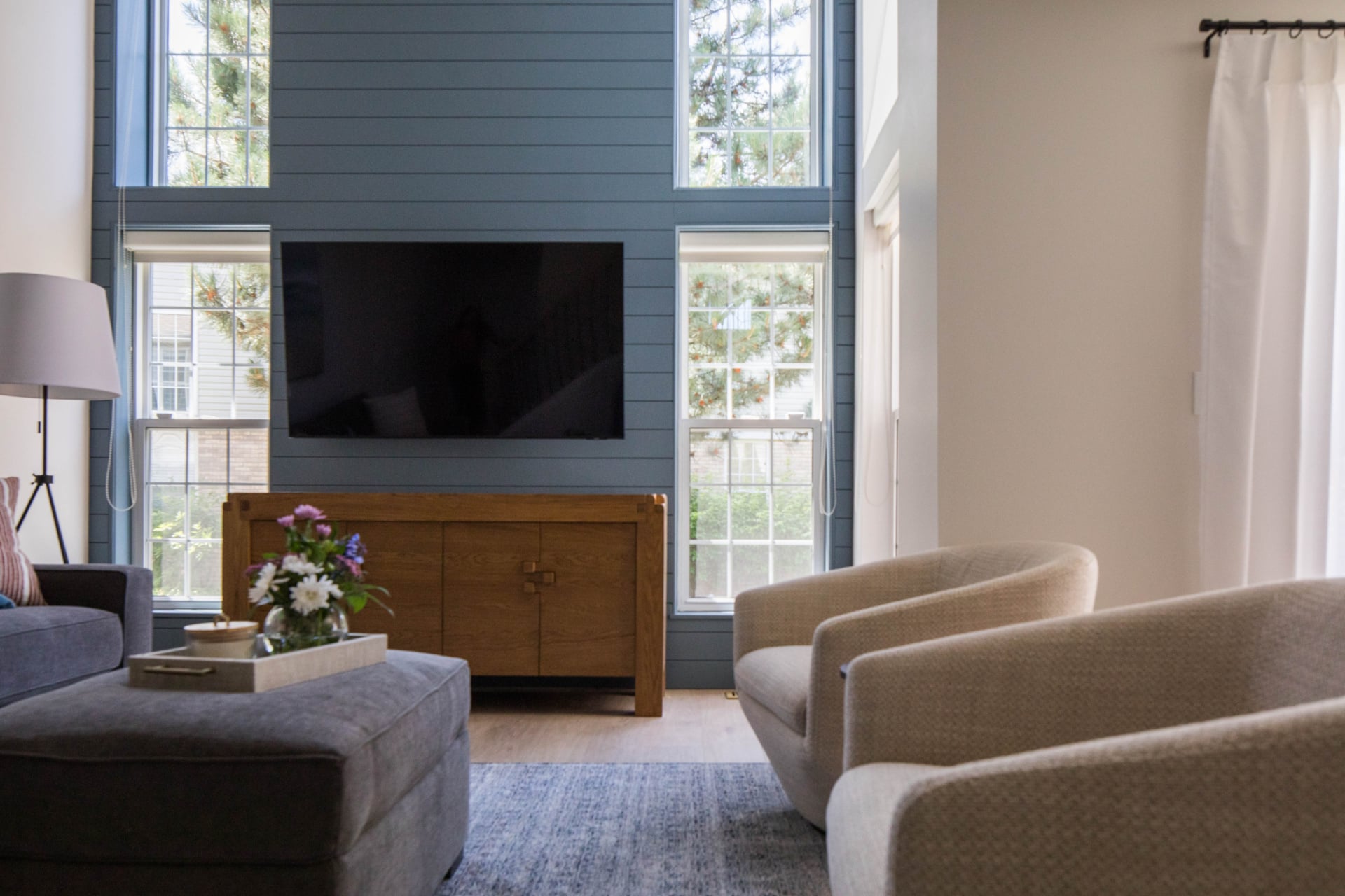 Modern living room with two beige armchairs, a gray ottoman, a wooden TV console, wall-mounted TV, and large windows with blue trim letting in natural light.