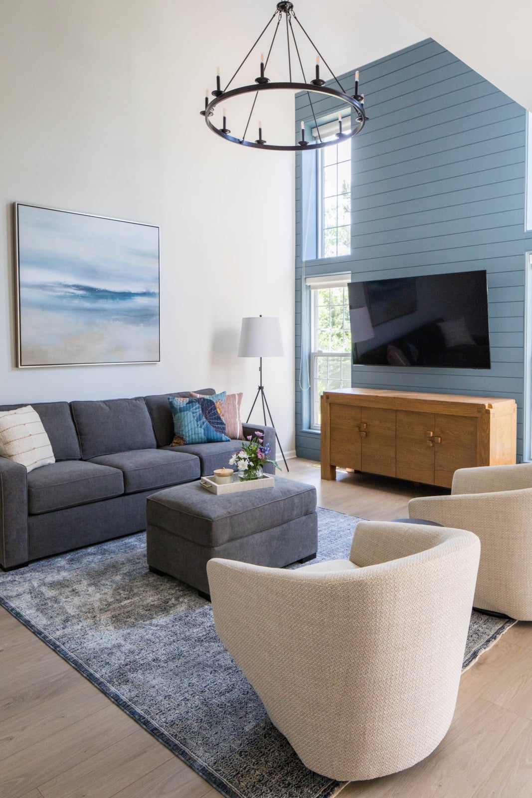 Modern living room with a gray sofa, two cream chairs, an ottoman, a wooden cabinet, wall-mounted TV, abstract art, and a large chandelier under high ceilings.