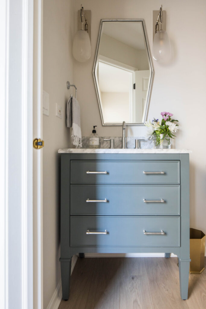 A modern bathroom vanity with a gray cabinet, marble countertop, rectangular sink, hexagonal mirror, wall sconces, and a vase of flowers on the counter.