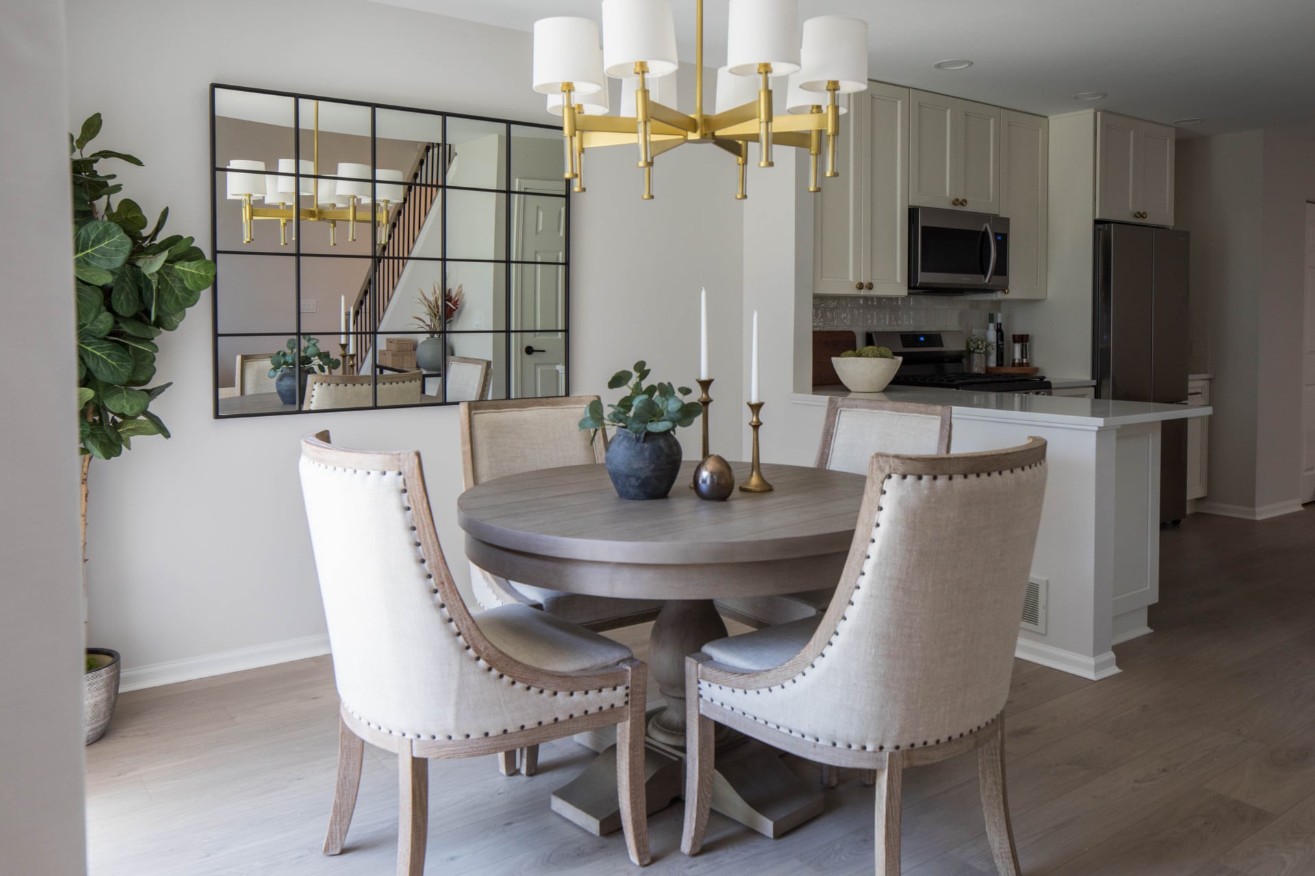 A modern dining area with a round wooden table, four upholstered chairs, decorative plants, a gold chandelier, and a kitchen in the background.