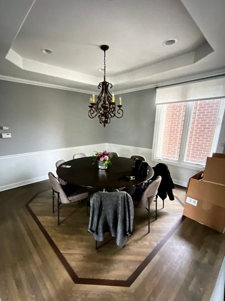 A round dining table with eight chairs, a chandelier overhead, a floral centerpiece, and boxes near the window in a gray-toned room with wood flooring.