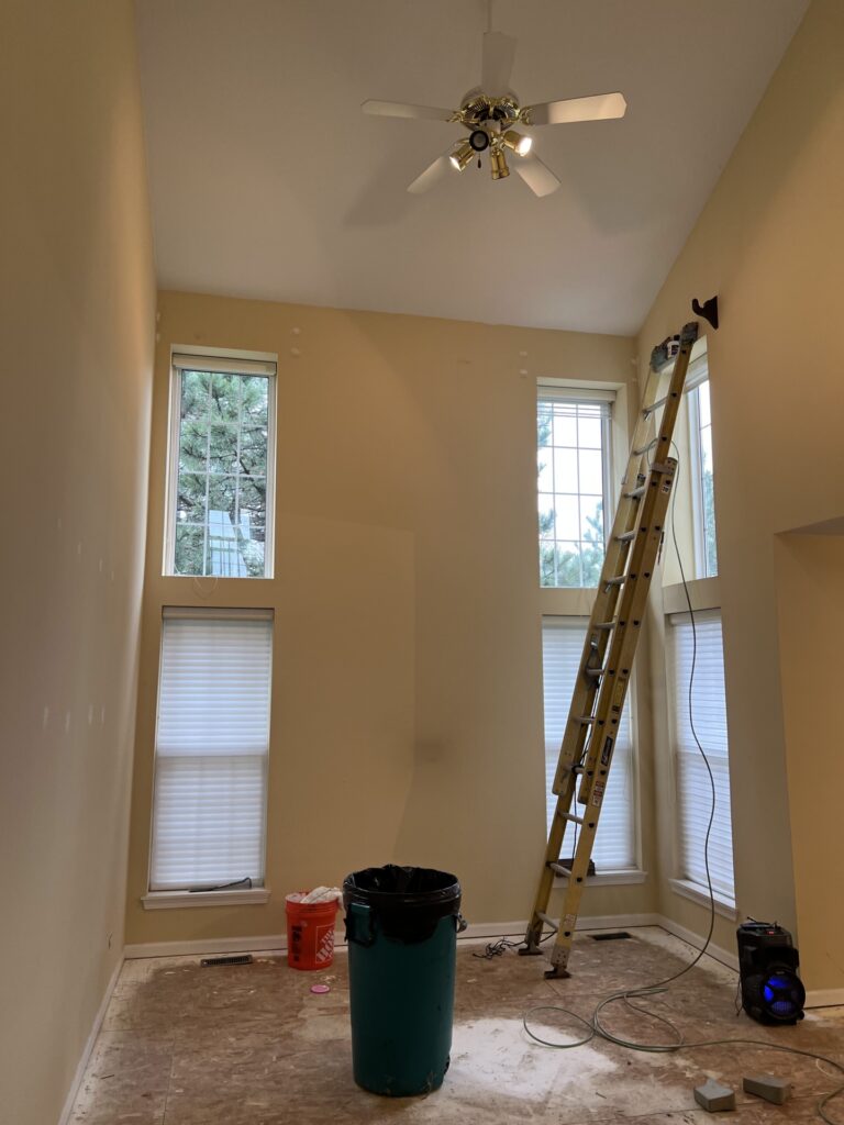Empty room with yellow walls, tall windows, a ceiling fan, an extension ladder, a trash bin, a red bucket, and some tools on the unfinished floor.